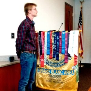 A young man wearing jeans, a plaid shirt and glasses stands next to a podium which is covered in ribbons and a flag representing Toastmasters International. 